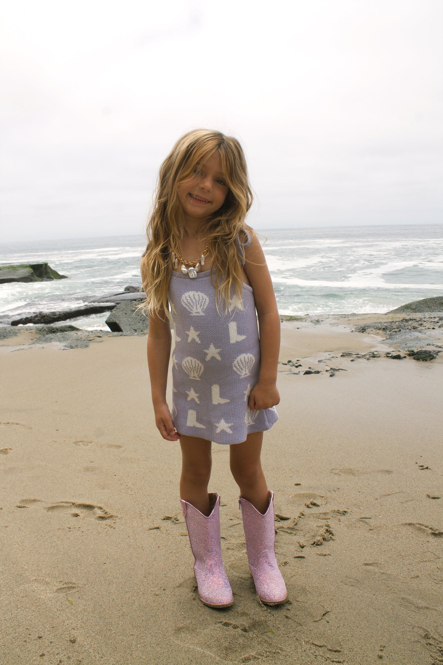 Toddler girl wearing purple Seaside Cowgirl knit dress with stars and boots on beach.