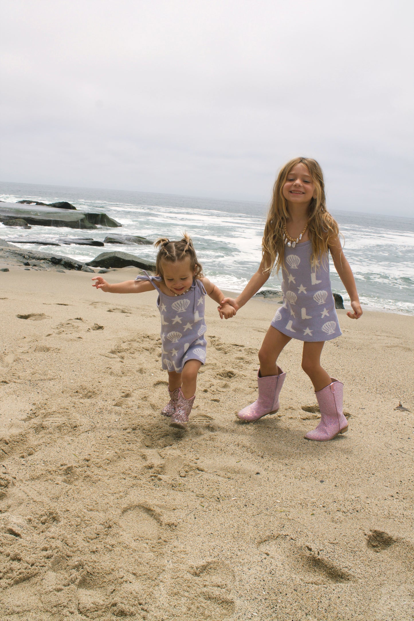 Toddler girl wearing purple Seaside Cowgirl knit dress with stars and boots on beach.