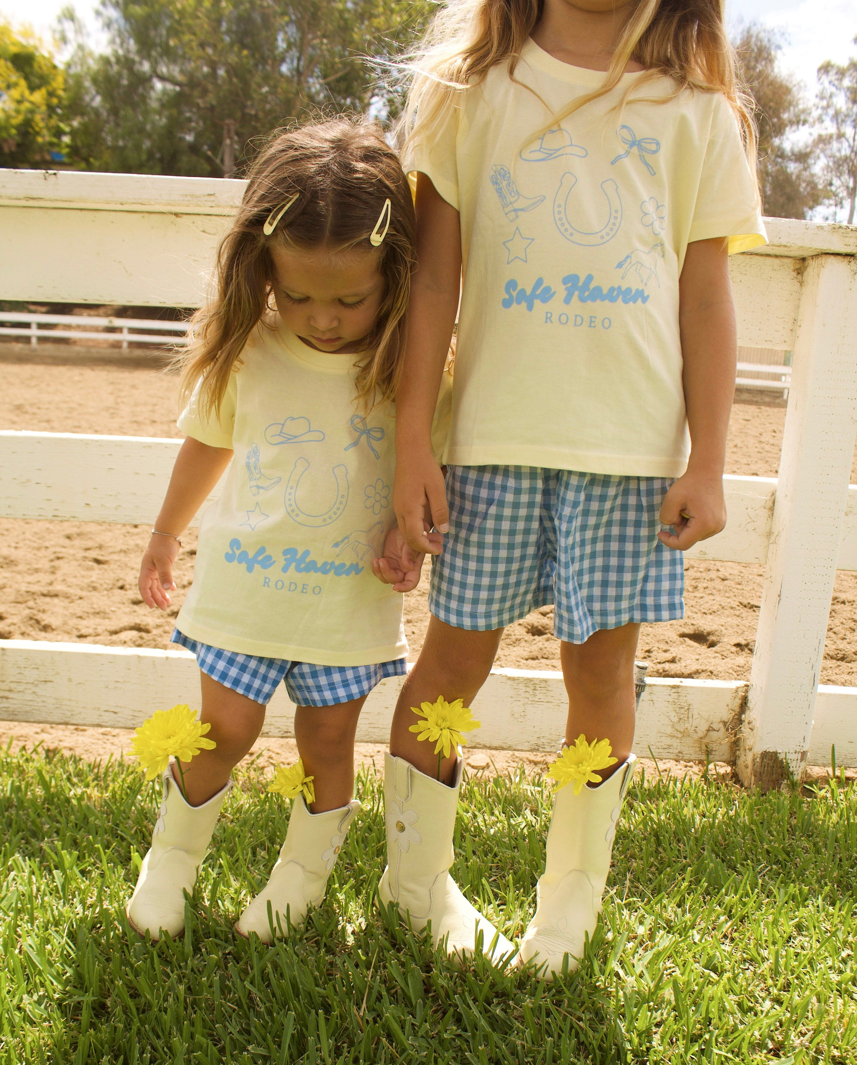 Toddler wearing a yellow “Safe Haven Rodeo” t-shirt with blue western illustrations, paired with blue and white gingham shorts and white cowboy boots, standing outdoors on green grass near a white fence.