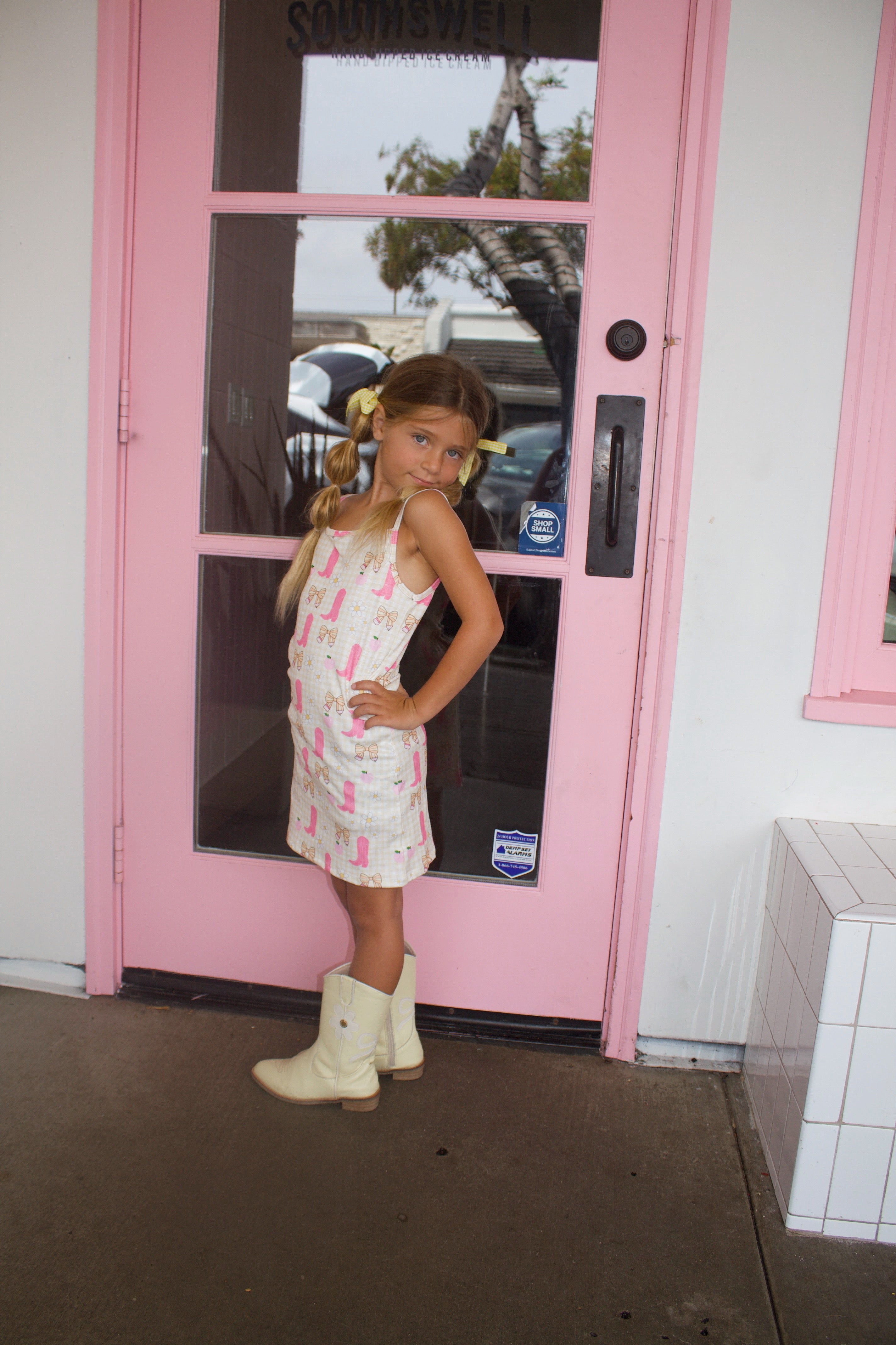 Smiling girl with pigtails and a yellow bow lying on a tiled bench in a pink and cream cowgirl-themed dress and boots in front of an ice cream shop.