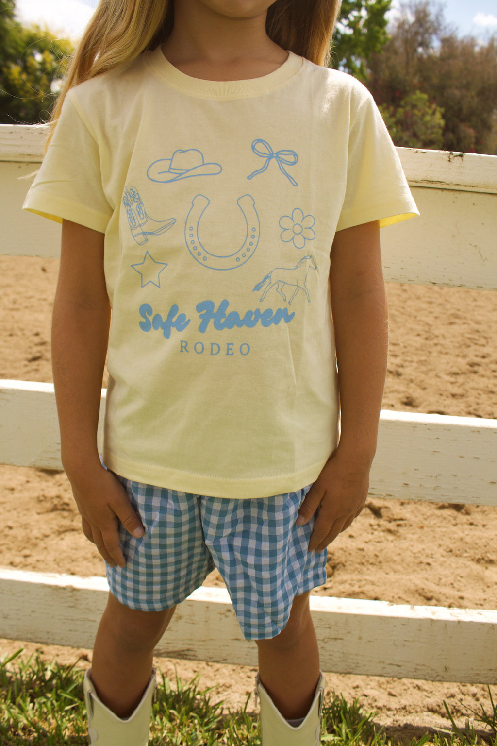 Toddler wearing a yellow “Safe Haven Rodeo” t-shirt with blue western illustrations, paired with blue and white gingham shorts and white cowboy boots, standing outdoors on green grass near a white fence.