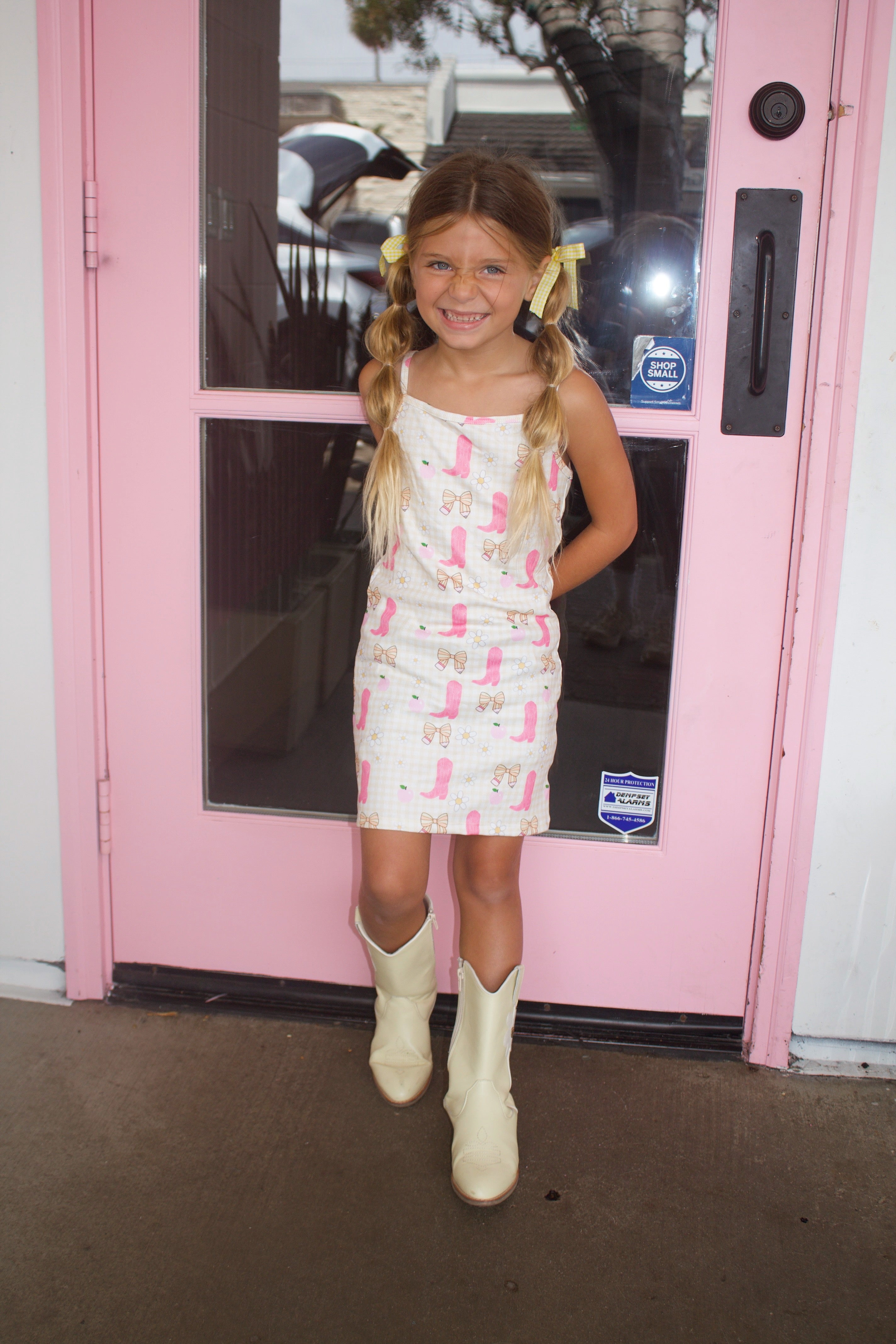 Smiling girl with pigtails and a yellow bow lying on a tiled bench in a pink and cream cowgirl-themed dress and boots in front of an ice cream shop.