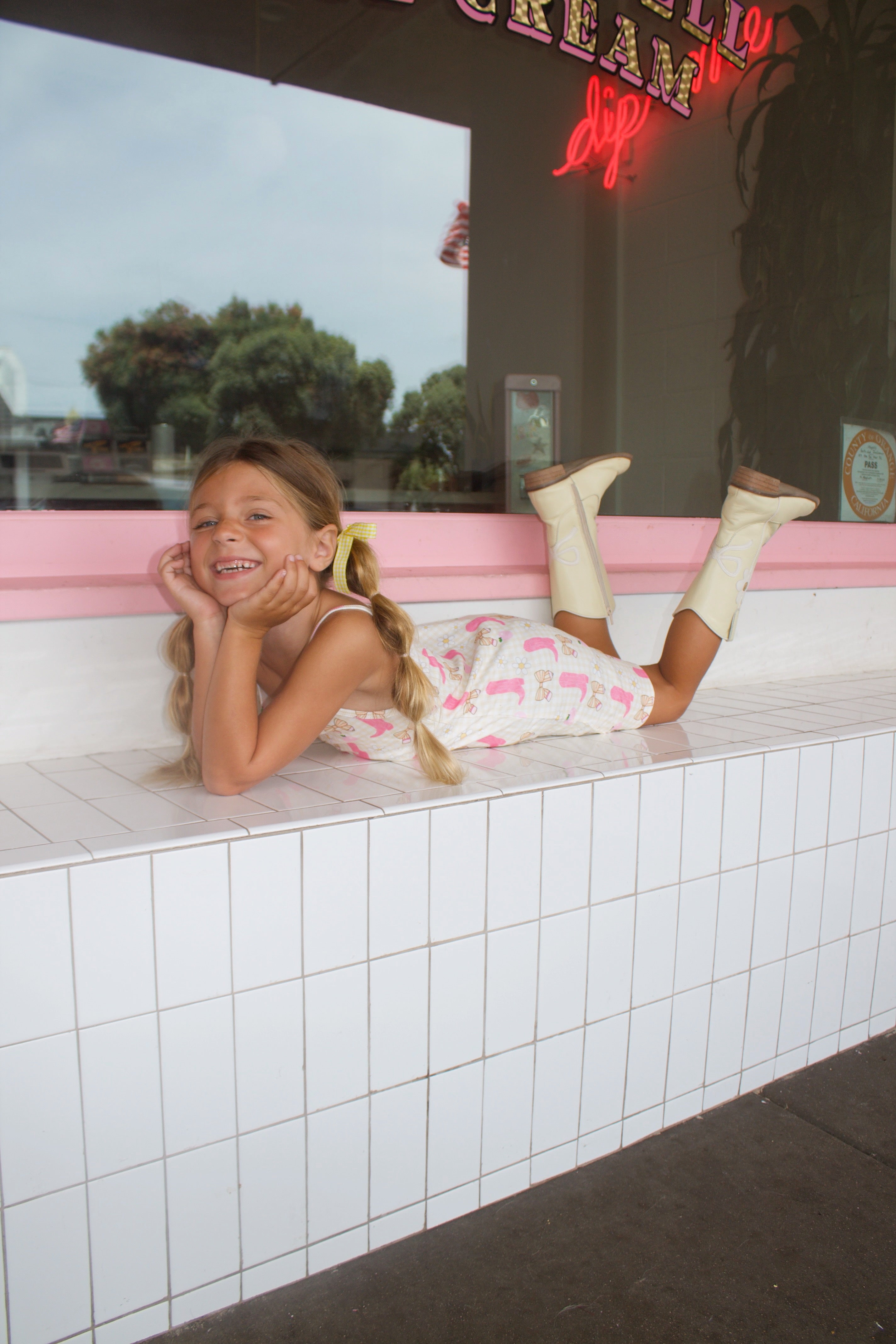 Little girl wearing a cream gingham dress with pink cowgirl boots, bows, daisies, and apple prints, paired with ivory western boots.