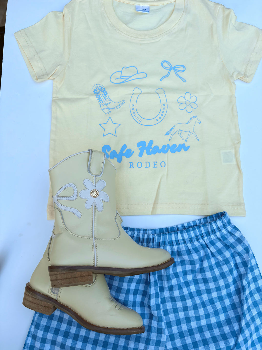 Toddler wearing a yellow “Safe Haven Rodeo” t-shirt with blue western illustrations, paired with blue and white gingham shorts and white cowboy boots, standing outdoors on green grass near a white fence.