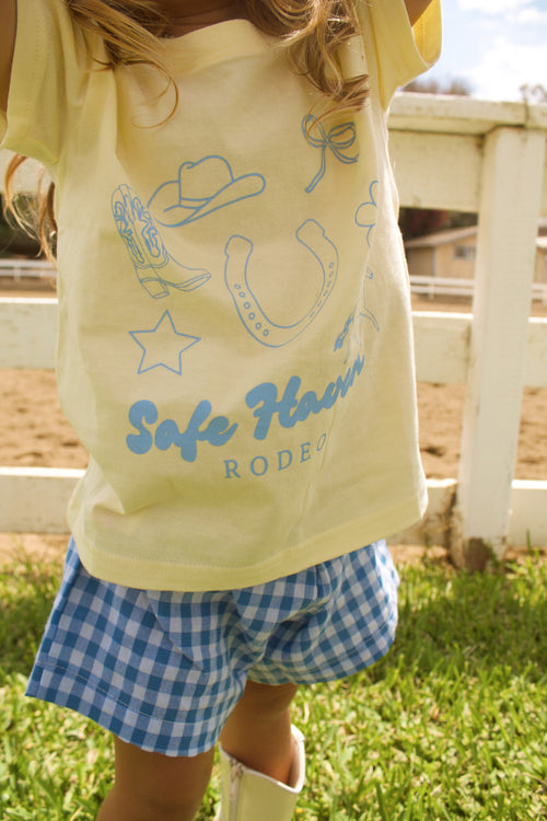 Toddler wearing a yellow “Safe Haven Rodeo” t-shirt with blue western illustrations, paired with blue and white gingham shorts and white cowboy boots, standing outdoors on green grass near a white fence.