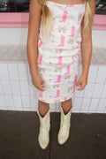 Little girl wearing a cream gingham dress with pink cowgirl boots, bows, daisies, and apple prints, paired with ivory western boots.