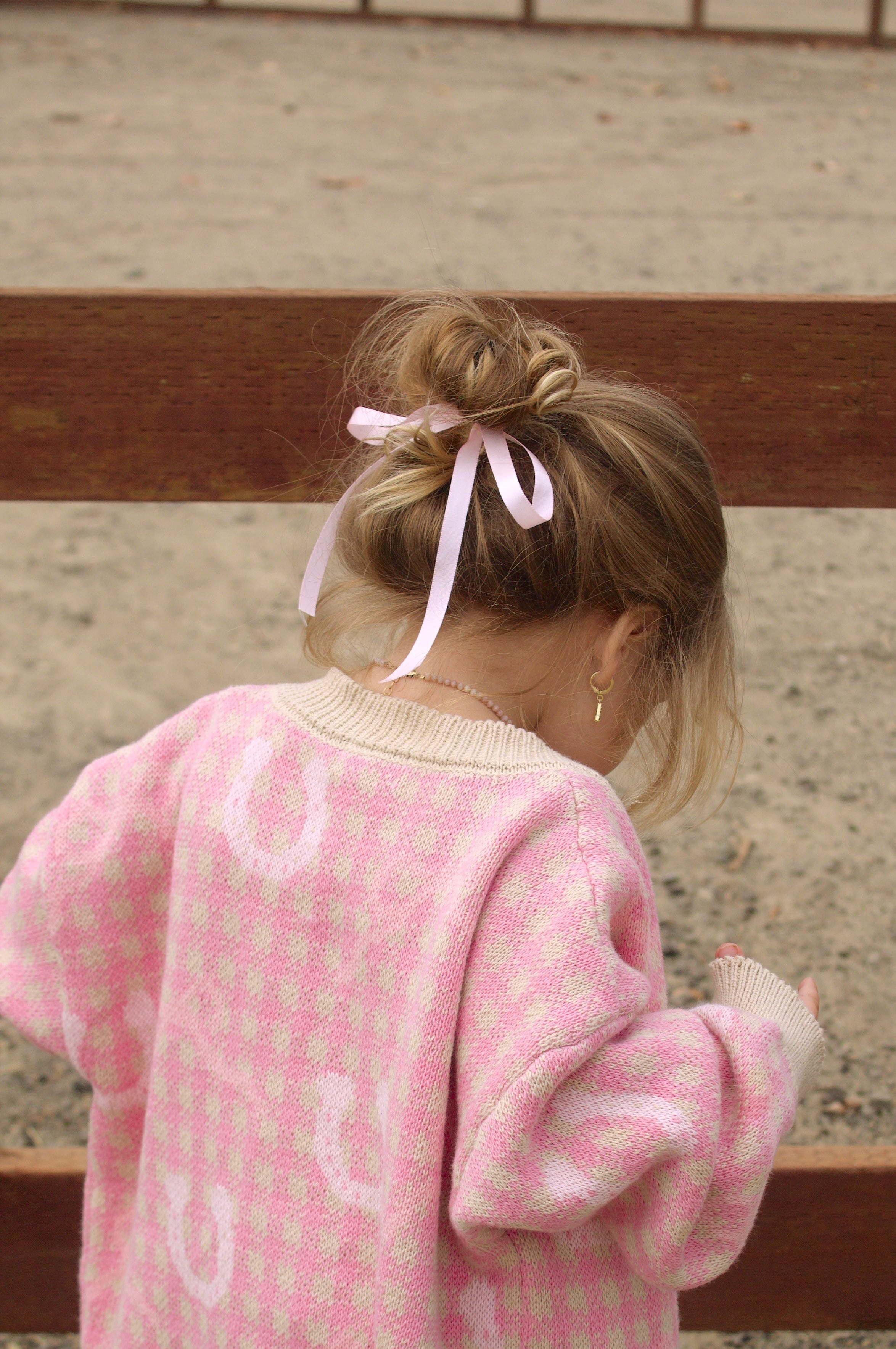 toddler girl wearing a pink cardigan with horseshoe and bow patterns, standing near a wooden fence.